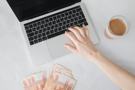 pexels-photo-6927397-6927397 Close-up of a hand on laptop keyboard with cash and coffee on table.